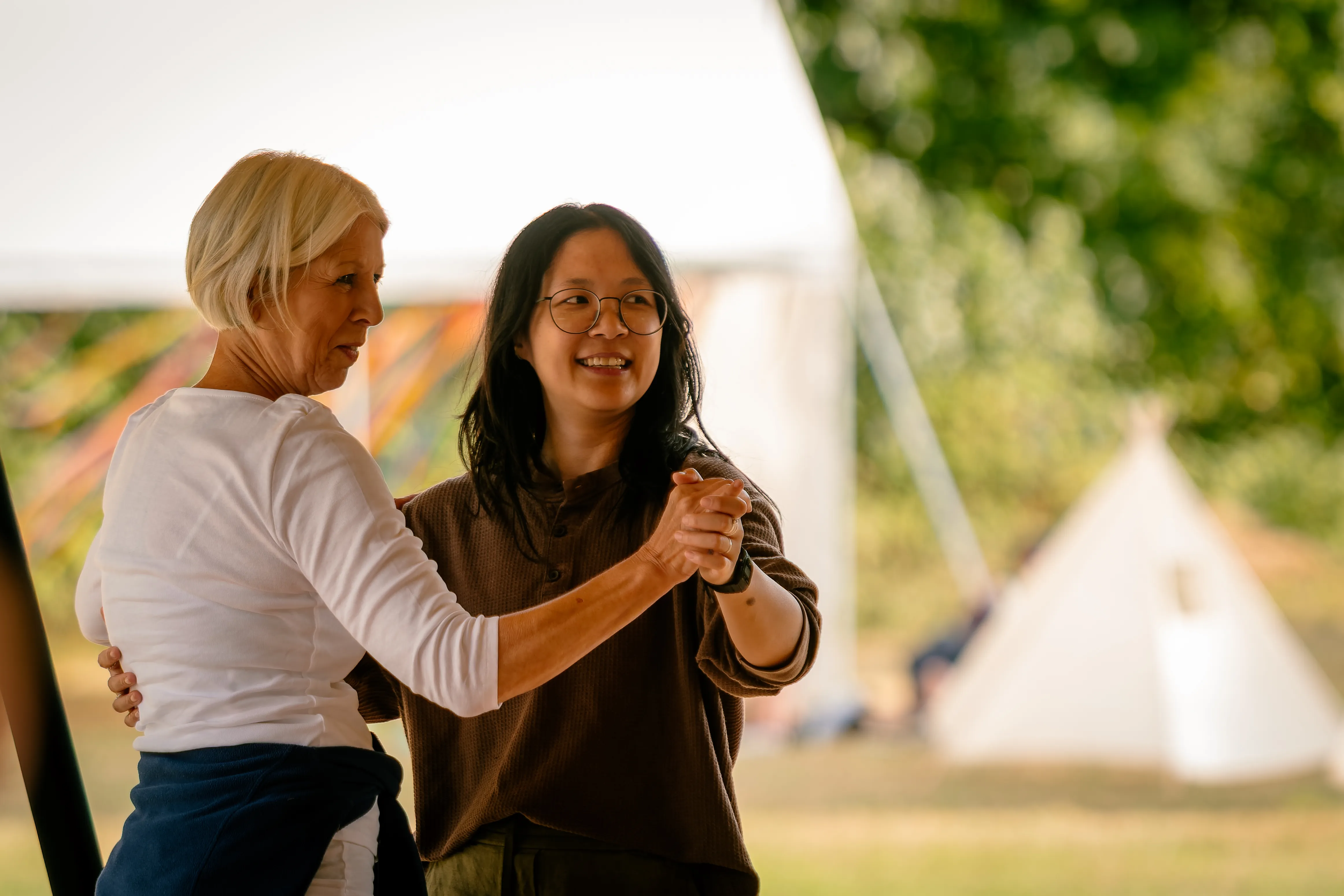 Two women in a gentle ballroom hold, one smiling at the other, a tipi in the background.