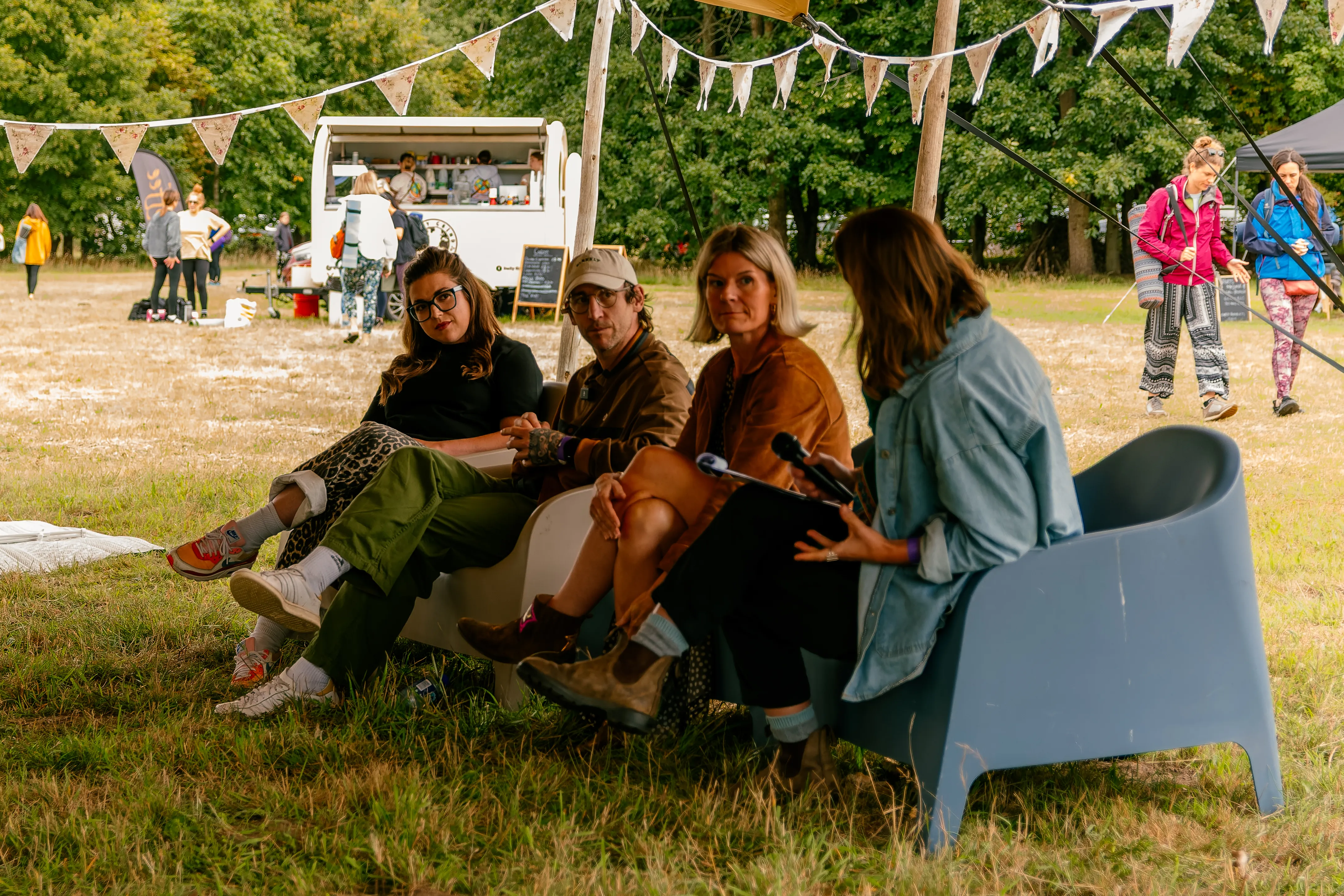 Four speakers in conversation on chairs under bunting, a coffee van and bunting-strung field behind them.