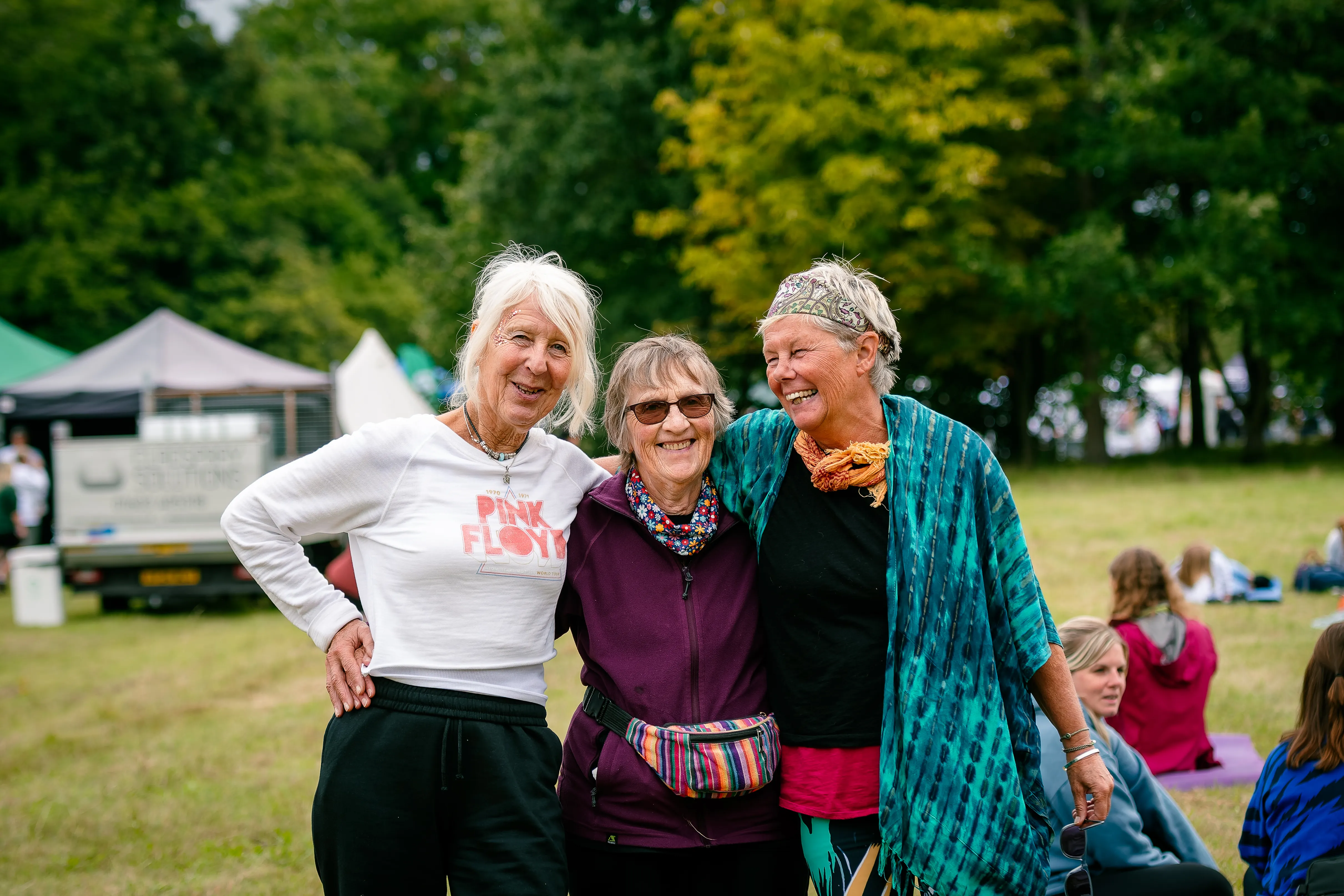 Three older women with their arms around each other, laughing together in the festival field.