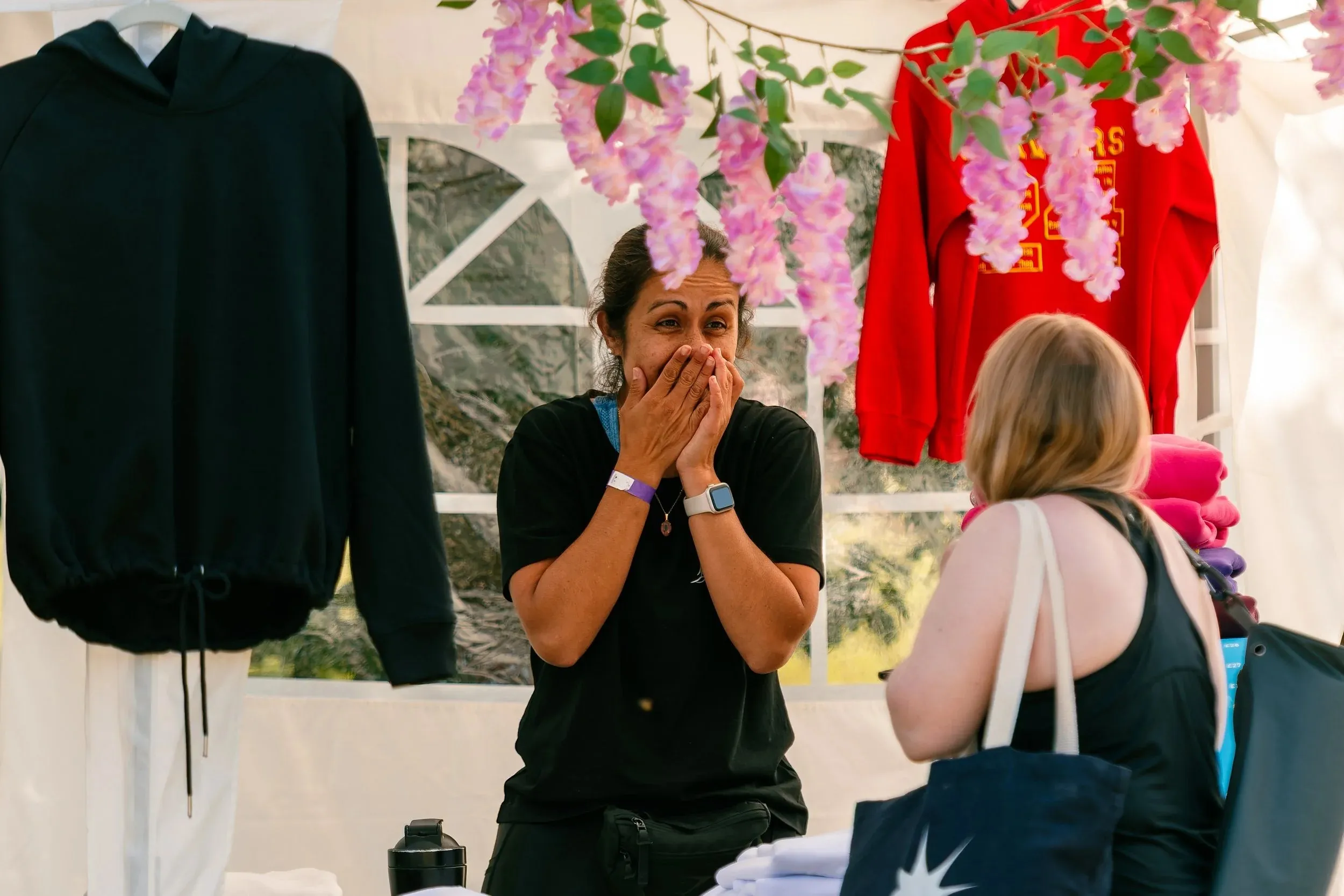 A woman covering her mouth in delight at a festival clothing stall hung with flowers.