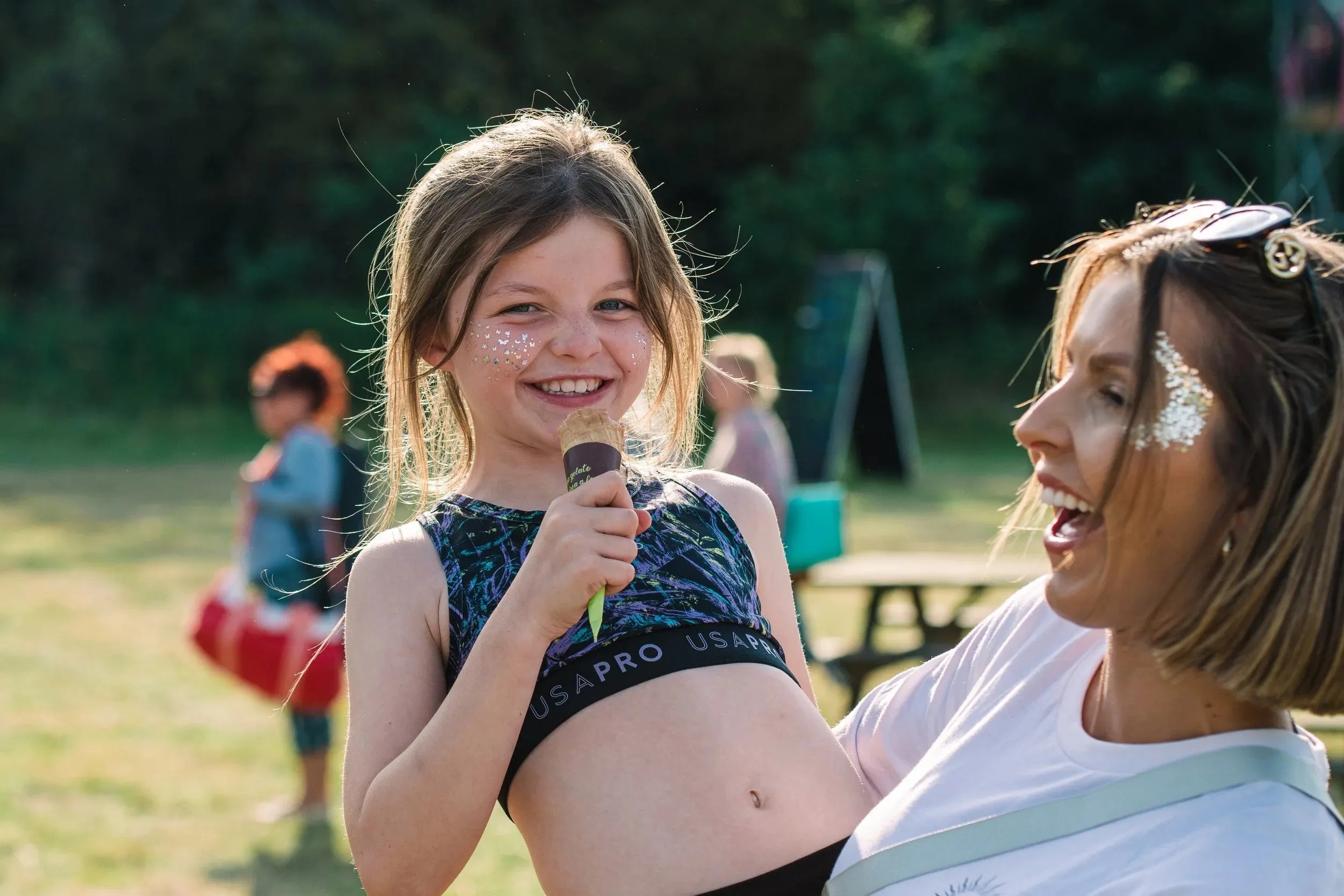 A young girl singing into a microphone beside a laughing woman with glitter on her face.